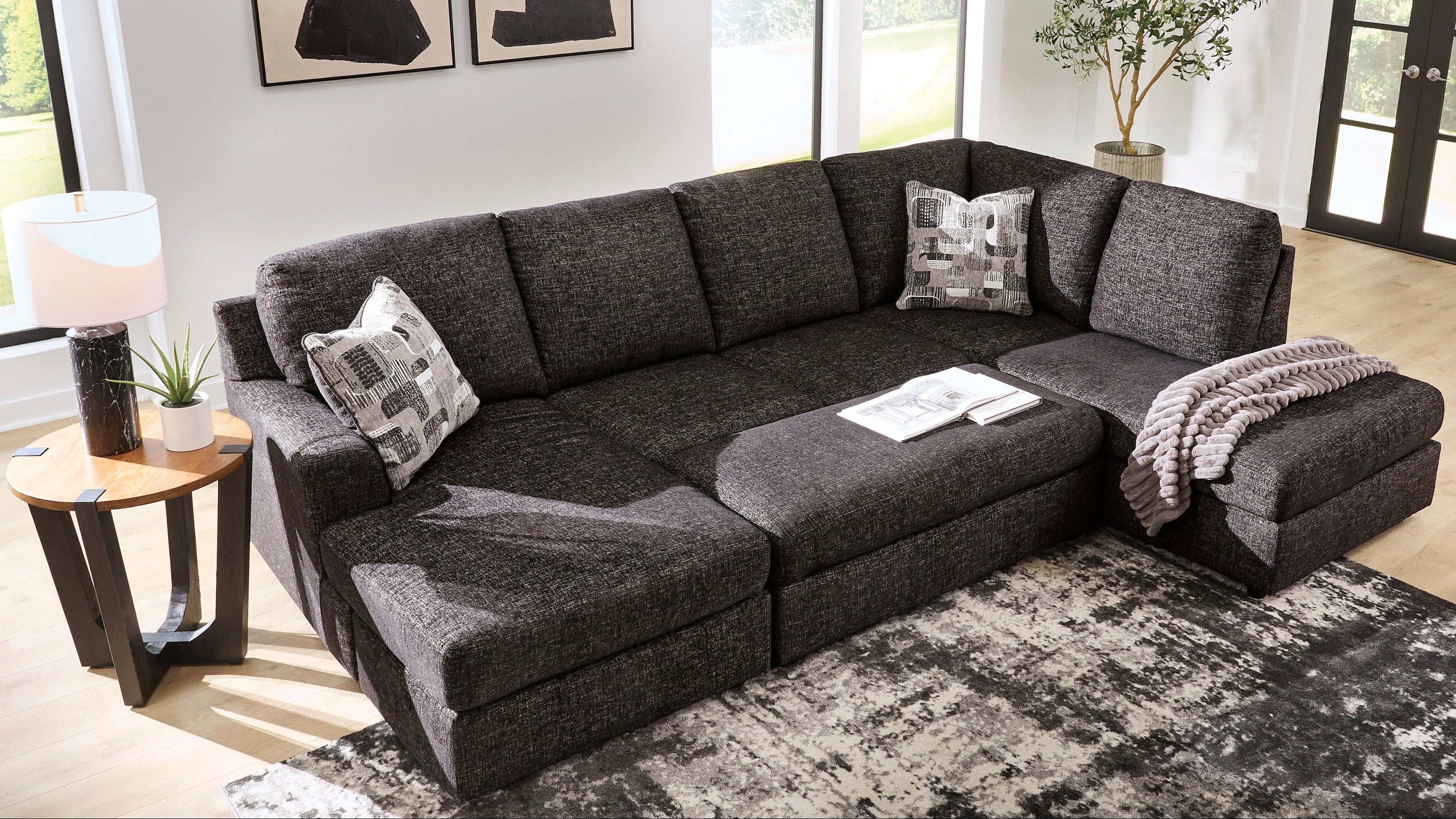 A dark gray fabric sectional sofa with a left-facing chaise lounge, styled in a bright modern living room with abstract wall art, a side table, and a patterned rug.